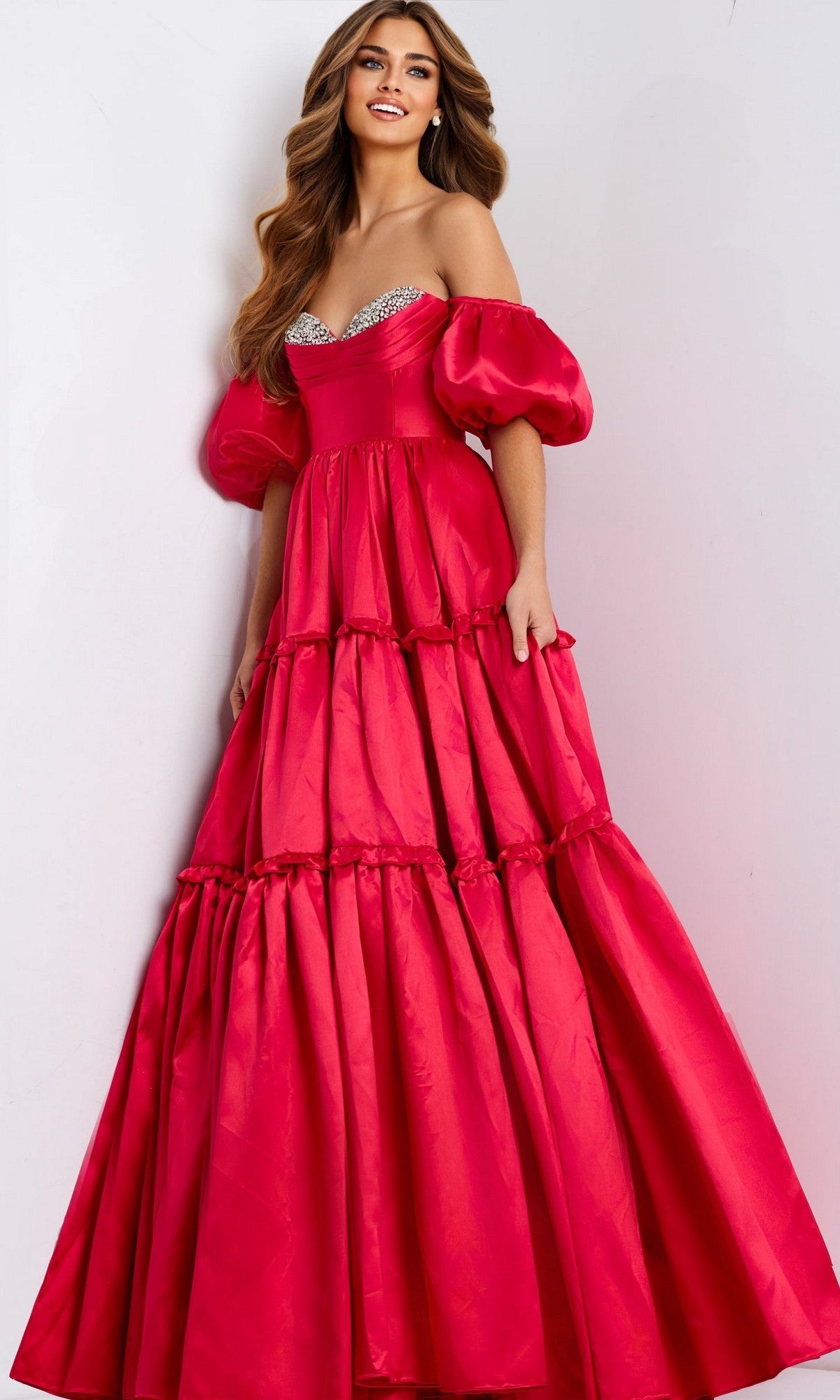 A woman radiates elegance in the striking Jovani 49055 long red prom dress, featuring off-the-shoulder satin, puff sleeves, a rhinestone-embellished bust, and a tiered skirt against a plain background.